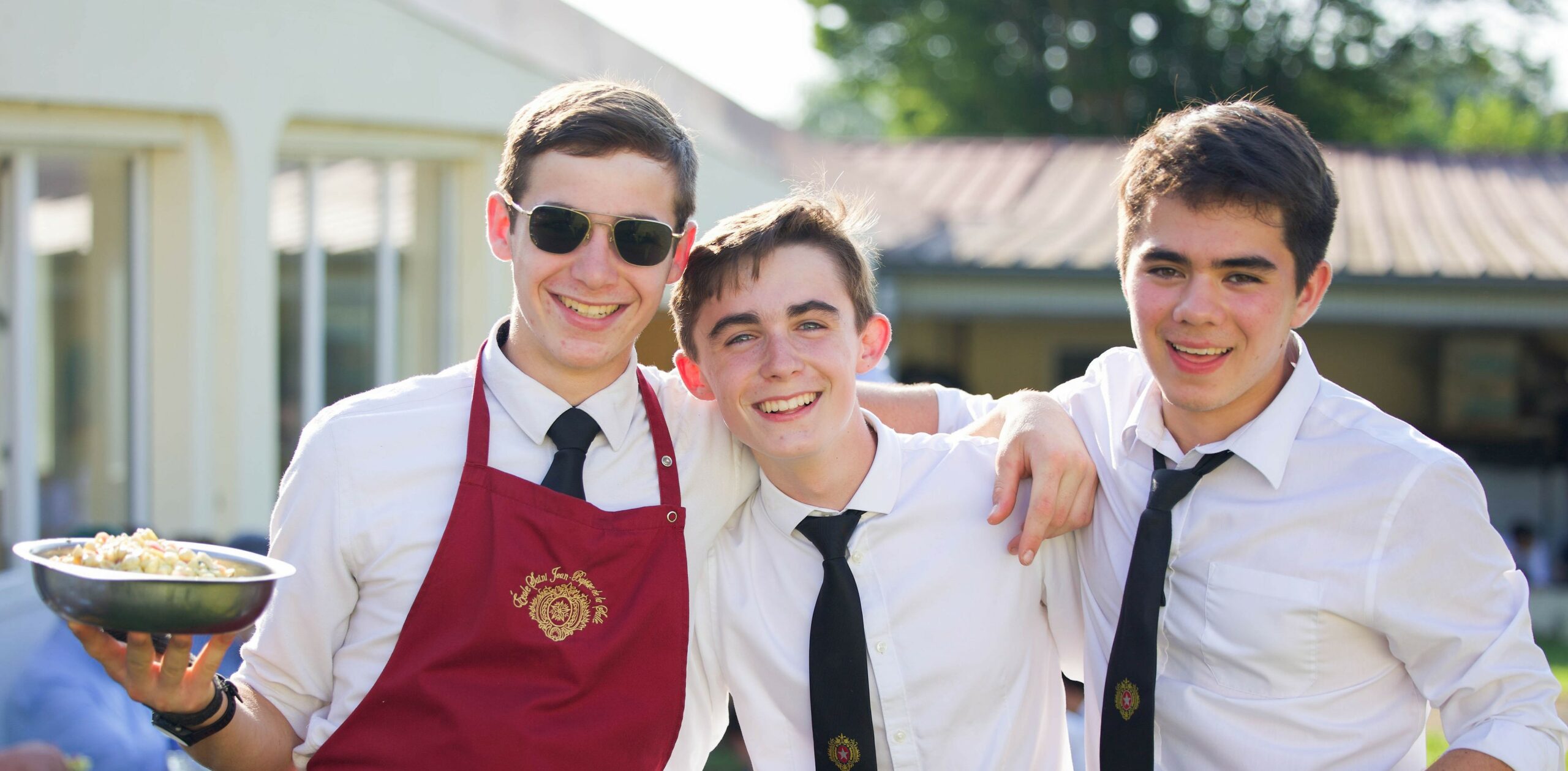 Trois élèves souriants lors du repas de fête, l’un en tablier de service